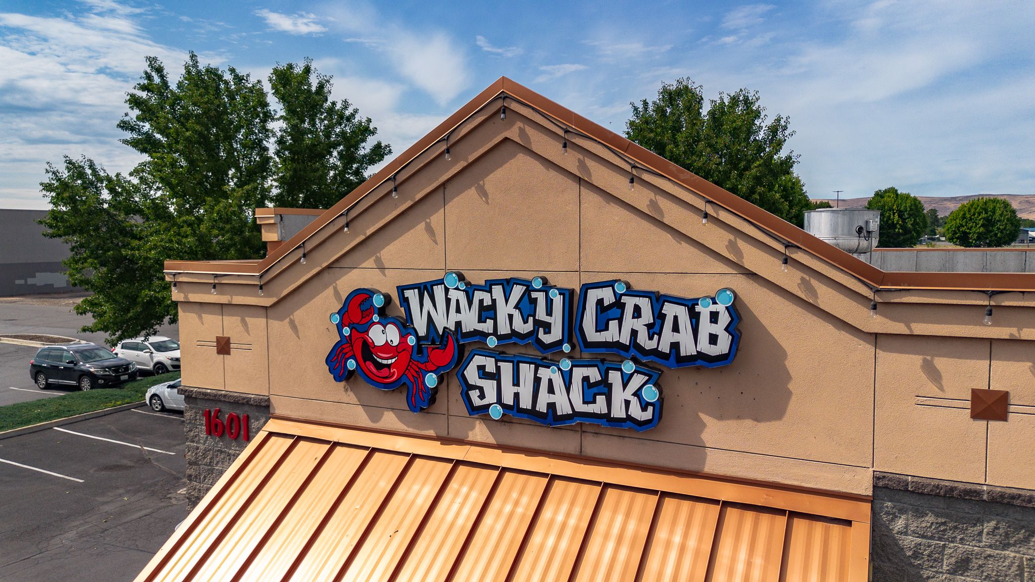 Drone-level close-up of the Wacky Crab Shack sign — the smiling red crab wordmark under the shack's orange-metal awning in Union Gap